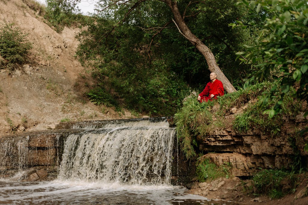 A monk in a red gown meditates by a serene waterfall in a tranquil outdoor setting.
